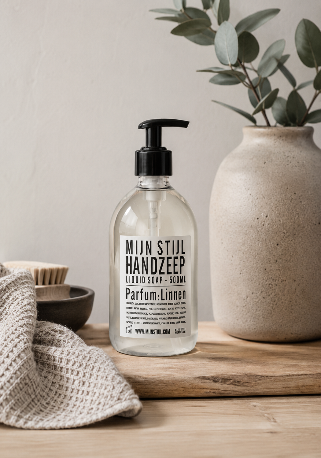Mijn Stijl handzeep Linnen (500 ml) in een transparante fles met zwart pompje, staand op een robuuste houten plank. Links op de voorgrond ligt een beige, geweven handdoek en daarachter staat een donker schaaltje met een houten afwasborstel. Rechts staat een grote, beige vaas van aardewerk met eucalyptustakken tegen een lichte wand.